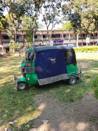 A green three-wheeled auto-rickshaw (CNG) is parked on a grassy and earthen patch in Shibpur, Bangladesh. The vehicle features a dark green canopy, a metallic front guard, and a dark blue side curtain with a gray bottom edge, displaying a gold "Style Gallery Silks & Sarees" emblem. In the background, a two-story building identified as "Shaheed Matiur Rahman College" by a sign in Bengali, presents a light-colored facade with multiple windows and red-brown trim along the upper floor. Tall deciduous trees with green leaves are densely distributed around and behind the college building. Green shrubs line the base of the college structure. The foreground consists of grass and exposed soil with scattered small rocks. The scene is outdoors under natural daylight; no people are visible.