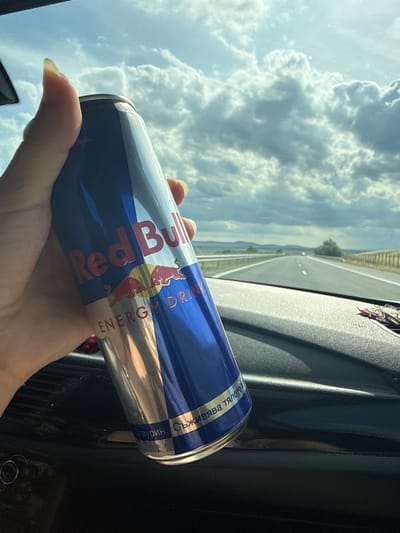 A hand holds a Red Bull energy drink can inside a vehicle. The can is blue and silver with the Red Bull logo visible. The background shows a highway stretching into the distance under a cloudy sky. The interior of the vehicle, including the dashboard, is visible in the foreground. The location is identified as Polsko Padarevo, Bulgaria.