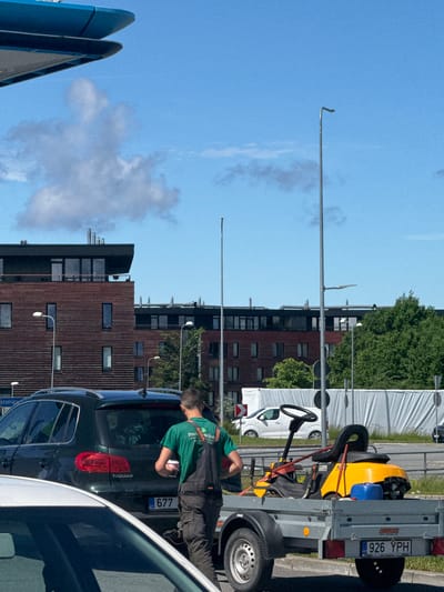 A person wearing a green t-shirt and dark overalls is standing next to a dark green Volkswagen Tiguan, holding a pink cup. The vehicle has the license plate "EST 677". Behind the car, a yellow and black riding lawnmower is loaded onto a silver trailer, which has the license plate "926 YPH". The trailer is attached to a vehicle that is not fully visible. In the background, there are modern apartment buildings with red brick facades and dark windows. A large white banner or tarp is visible behind the trailer. The scene appears to be a street or parking area, with streetlights and trees present. The sky is blue with scattered clouds. The location is Tiskre, Estonia.