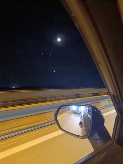 This media file captures a nighttime view from inside a moving vehicle, likely on a highway or bridge, in Surakhany Raion, Azerbaijan. The scene is dominated by the dark night sky, illuminated by a bright moon and what appear to be two other distant lights or celestial bodies.

The foreground features the interior of the car, with the right side door frame and a side-view mirror visible. The mirror reflects the road and headlights of other vehicles, suggesting the car is in motion. Streaks of light blur across the image, indicating a fast shutter speed and the movement of the car.

The middle ground shows a highway or bridge with yellow guardrails and striped lanes. The road surface is brightly lit, casting a warm glow that contrasts with the dark sky. The distant horizon is obscured by darkness, but faint lights can be seen, suggesting a built-up area or city lights in the distance.

The overall mood is one of travel and observation at night. The bright moon and the illuminated road create a sense of calm, despite the implied speed of the vehicle. The location context of Surakhany Raion, Azerbaijan, places the scene in a specific geographical area, adding to its factual description. There is no visible text in the image.