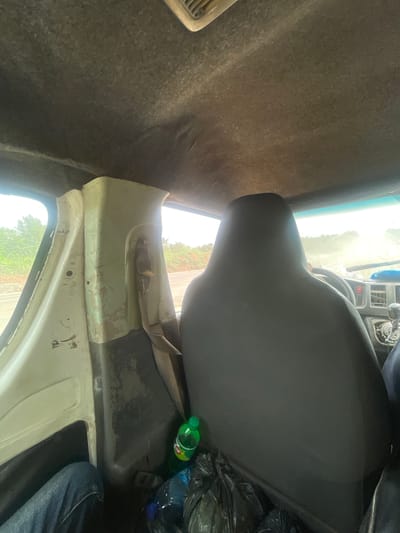 The image captures the interior of a vehicle, likely a truck, in Ago-Abo, Nigeria. The perspective is from the rear passenger seat, looking forward and to the left. The ceiling of the vehicle appears worn and stained, with visible air vents. To the left, a white interior paneling shows signs of wear, with peeling paint and exposed screw holes. A brown seatbelt is attached to this panel. Outside the vehicle, seen through the windows, is a rural landscape with greenery and a dusty road.
In the foreground, a dark gray seat is prominent, occupying the right side of the frame. Next to it, on the floor, are several black plastic trash bags and a green bottle of 7UP. The driver's side dashboard is partially visible, including air vents and part of the steering wheel, suggesting the vehicle is in motion or recently stopped. The overall impression is one of a utilitarian vehicle used for transport in a rural setting.