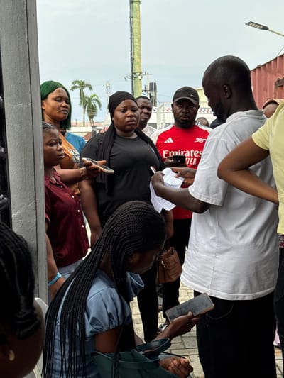 A group of approximately 10 people, mostly of African descent, are gathered outdoors in Ojomu, Nigeria. The setting appears to be an urban environment, indicated by buildings, palm trees, and overhead utility lines. A few individuals are looking at phones and papers, suggesting a potential transaction or consultation. Several people are standing close together. The image composition is a low-angle shot.