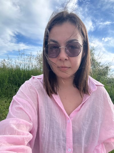 A young woman with shoulder-length brown hair and purple tinted sunglasses is captured in a selfie outdoors. She is wearing a crinkled, light pink button-up shirt. The background consists of tall green grass, sparse dry reeds, and a sky with scattered white clouds. The scene suggests a natural, possibly rural or park-like setting, consistent with locations found in Ozyorny, Russia.