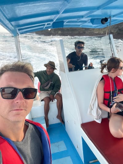 A selfie on a blue boat with five people visible. The photographer wears a life vest and sunglasses. A woman sits on a red bench with a white towel and red life vest, holding a phone. A driver wearing sunglasses controls the boat. Another man in a hat and jacket sits with legs outstretched. Ocean waves are visible outside the boat.