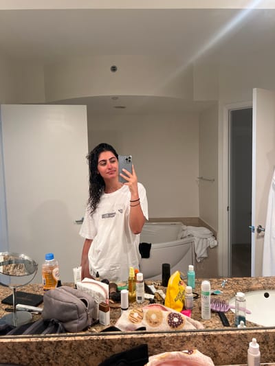 The image shows a woman standing in front of a bathroom mirror in a hotel room. She is wearing a white t-shirt and has her hair pulled back from her face. The woman is holding a smartphone and is looking at herself in the mirror. There are various toiletries on the counter behind the woman, including bottles of shampoo, conditioner, and lotion. There is also a washcloth with a donut pattern on it. The bathroom is tiled and has a white bathtub. The location is a hotel room in Miami Beach, Florida, United States. The coordinates, latitude 25.773 and longitude -80.141, place the scene in the heart of Miami Beach, known for its luxurious hotels and beaches.