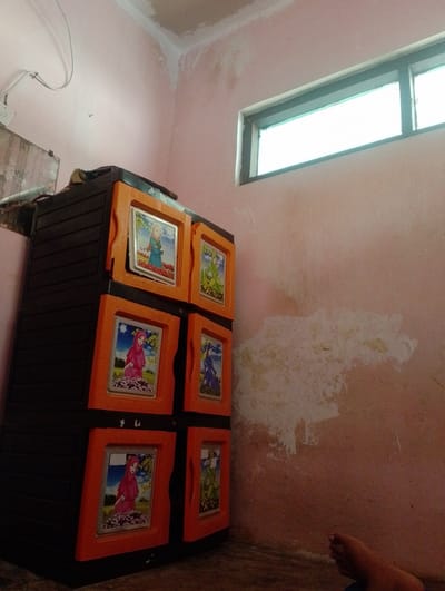 In the corner of a room with pink walls, a black and orange plastic cabinet is visible. The cabinet has four doors, each decorated with an image of a person wearing a hijab in a garden setting. The wall has patches of peeling paint. A narrow window with a light green tint is positioned high on the wall. A coiled white electrical wire hangs from the ceiling. The floor is made of brown tile. A bare foot is visible in the lower right corner of the image. The overall scene appears to be an interior residential space in Sukamanah, Indonesia.