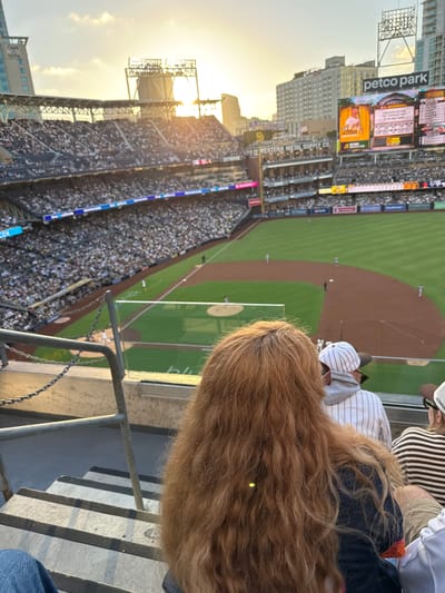 This is a horizontal, eye-level, medium shot taken from stadium seating at Petco Park in San Diego, California, during a baseball game. The sun is setting, casting a warm glow over the stadium. In the foreground, the back of a person with long, wavy, red hair fills the right side of the frame. To the left of this person, partially visible, is another spectator wearing a white and grey pinstripe baseball cap and hoodie. Further to the left, a portion of the stadium seating is visible, packed with spectators. The baseball field is in the midground, with players on the infield. The background shows the cityscape of San Diego, including tall buildings and the "Petco Park" sign. The stadium is illuminated by bright lights, suggesting the game is ongoing.