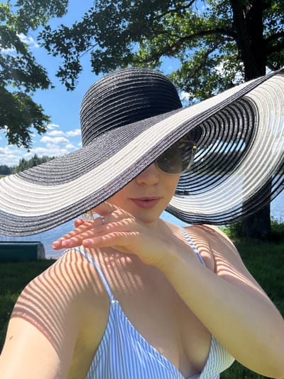 A woman wearing a wide-brimmed black and white striped hat and sunglasses is pictured outdoors on a sunny day. She is wearing a blue and white striped swimsuit. Her left hand is raised, with her fingers partially covering her face. Sunlight casts striped shadows across her arm and shoulder. In the background, there is a body of water, green grass, trees, and a blue sky with some clouds. The location is not specified to be in Lendži, Latvia, or any other city.
