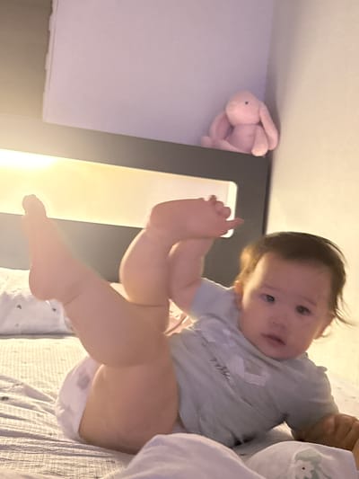 A baby is lying on a bed in a room. The baby is wearing a light blue shirt and a diaper. The baby's legs are in the air, and its feet are visible. A pink stuffed bunny is on a shelf in the background. The room is dimly lit, suggesting it might be nighttime or the blinds are drawn. The baby appears to be looking at the camera with a curious expression. The overall mood of the image is relaxed and intimate, capturing a moment of playfulness with a loved one. The location context is Gwangju, South Korea. There is no visible text in the image.
