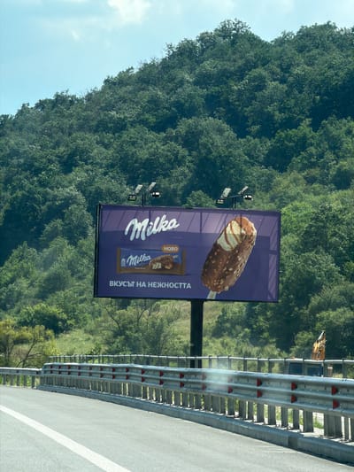 The image shows a billboard for Milka ice cream located in Churek, Bulgaria. The billboard features a large image of a chocolate-covered ice cream bar with caramel swirls and chopped nuts, along with the Milka logo and text that reads "HOBO" and "ВКУСЪТ НА НЕЖНОСТТА" (The taste of tenderness). The billboard is placed on the side of a road with a highway guardrail in the foreground. The background consists of a lush green hillside covered in trees and a bright blue sky. The scene appears to be a sunny day.