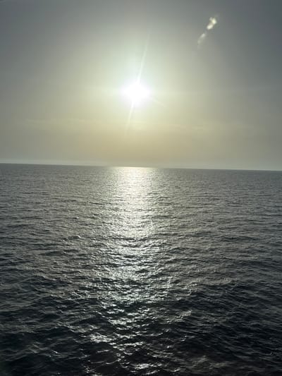 The image displays a wide, unobstructed view of the Mediterranean Sea under a bright, hazy sun. The sun is positioned in the upper center of the frame, casting a strong glare and a shimmering reflection across the water's surface. The sea itself is calm, with gentle ripples creating a textured pattern of light and shadow. The horizon line is clear, separating the vast expanse of water from the pale, diffused sky. There are no discernable landmasses or man-made structures visible in this depiction of the sea. The location is identified as Ain El Turk, Algeria.