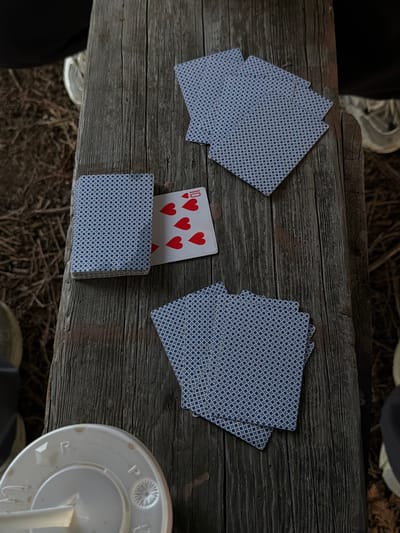 An overhead, slightly angled shot captures a rustic wooden picnic table with playing cards scattered across its surface. The table is weathered, with prominent grain lines and some discoloration.
On the table, there are several fanned-out piles of playing cards, all featuring a blue patterned back. One single card is visible face-up, revealing a "10" of hearts, with five heart symbols depicted. Beside the face-up card, a deck of cards is neatly stacked, also showing the blue patterned back.
To the lower left of the image, a white plastic cup lid with some embossed lettering is partially visible. The lid has embossed letters that appear to be "R" and "P" among other designs.
The background of the image shows a rough, ground cover of dry leaves and twigs, suggesting an outdoor setting. There are also glimpses of footwear, including a white sneaker on the left and a dark shoe with white laces on the right, indicating the presence of at least two individuals. The overall atmosphere is casual, possibly a game being played outdoors.
The scene is located on a wooden picnic table within the city of New Westminster, Canada.