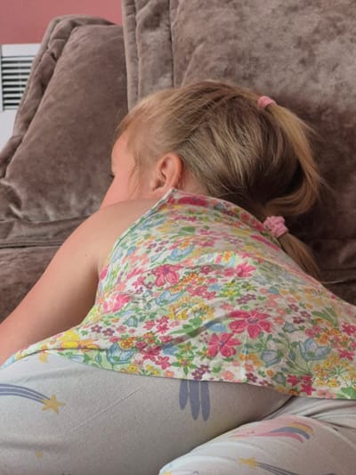 The image shows a close-up of a young child, likely a girl, lying down on a soft, brown, textured surface, possibly a couch or a bed. The child is facing away from the camera, with their head turned to the left. Their hair is blonde and pulled back in a ponytail with a small pink hair tie. They are wearing a colorful floral-print top with pink, yellow, blue, and green flowers. To the right of the floral top, the child is wearing light-colored pants with a pattern that includes shooting stars and rainbow-like arcs.
The setting appears to be indoors, possibly a home in Švenčionys, Lithuania. The lighting suggests it could be daytime, though there are no clear indicators of the weather or specific time of day. The child's posture and the overall scene evoke a sense of restfulness or sleep. No text is visible in the image.