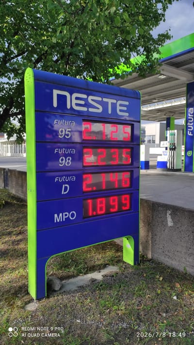This is a vertical, eye-level shot of a Neste gas station price display in Iisalmi, Finland. The display features four columns showing fuel prices. From left to right, the prices are: 1.729 for Futura 95, 1.729 for Futura 98, 1.619 for Futura D, and 1.669 for MPO. The "NESTE" logo is prominently displayed on the left side of the sign. Behind the price display, the canopy of the gas station is visible with green and blue elements, along with gas pumps. The scene is outdoors, with a tree on the left providing shade and greenery. The ground is a mix of pavement, gravel, and grass. The timestamp in the lower right corner indicates the photo was taken on July 8, 2026, at 18:49. The lighting suggests it is late afternoon or early evening. There are no people visible in the frame. The sky is overcast. The text "FUTURA" is visible at the top of the gas station canopy in Finnish.