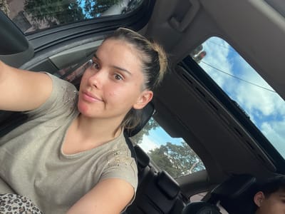 A selfie, taken from inside a car, shows a woman with light skin and brown eyes. She is wearing a beige shirt with sparkly embellishments. The car's sunroof is open, revealing a view of the sky and trees. The photograph was taken during daytime. It's unknown if this photograph was taken in San Isidro, Argentina.