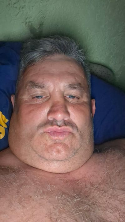 An extreme close-up selfie shows the face of a middle-aged man with grey hair and a hairy chest lying down on a pillow. The man is looking directly at the camera with his lips pursed. He has a double chin and his skin appears reddish. He is wearing a blue pillow with a yellow design on his left. The background is a textured green wall. The scene is indoors, likely in a bedroom. There is no information that can identify the location as Voinova, Moldova.