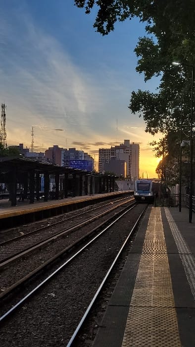 A train approaches along tracks under a vibrant sunset sky. Tall buildings form a backdrop, while leafy trees frame the left side of the scene. The train is simple in design, white and blue, nearing the perspective point. A platform with textured tiles runs along the right, beside the tracks.