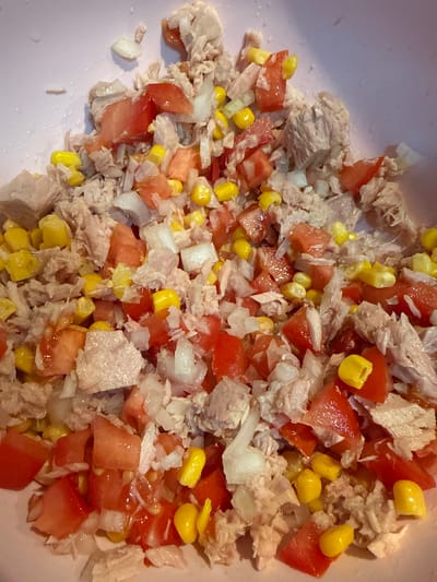 A close-up, overhead shot shows a bowl filled with a mixture of shredded tuna, diced tomatoes, finely chopped white onions, and bright yellow corn kernels. The ingredients are muddled together, indicating they are being prepared or have just been mixed. The lighting suggests an indoor setting, possibly a kitchen, with a soft light illuminating the food. The colors are vibrant, with the red of the tomatoes, yellow of the corn, and the pale pinkish-white of the tuna and onions creating a visually appealing dish. There are no people or explicit text visible in the image. The scene depicts the preparation of a simple, fresh salad or filling.