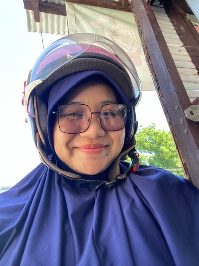 A selfie taken outdoors shows a young woman wearing a blue hijab and a pink and gray helmet with a clear visor. She has on black-framed glasses and is smiling. The background consists of a corrugated metal roof, wooden beams, and green trees under a bright sky. The image appears to be taken in Sungai Paring, Indonesia.
