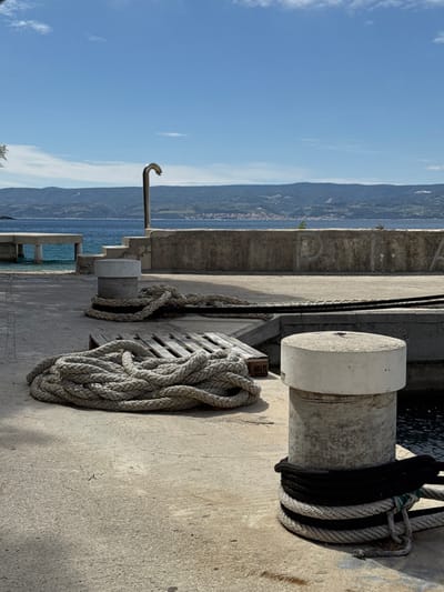 The image shows a concrete pier with two mooring bollards and a pile of thick rope lying on the ground. The bollards are made of concrete and are cylindrical in shape. The rope is made of natural fibers and is coiled into a neat pile. There is a wooden pallet underneath the rope. In the background, there is a concrete wall and a blue sea. The image is taken from a slightly elevated angle, and the pier is empty except for the rope and the bollards. The coordinates provided place the location in Dugi Rat, Croatia, overlooking the harbor. The scene appears calm and serene. The concrete wall has some graffiti markings on it. The water level is visible behind the pier wall. There is a silver shower head on the wall in the background. The image was taken on a sunny day.
