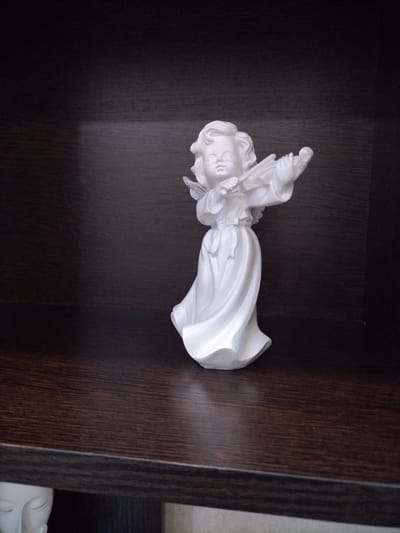 A white ceramic figurine of an angel is positioned on a dark wooden shelf. The angel is depicted playing a violin, with its left hand on the fingerboard and its right hand holding the bow. It wears a flowing, draped robe that covers its legs and feet, and delicate wings extend from its back. The figurine has a youthful appearance with curly hair and a serene expression. The background consists of a dark, plain surface, likely the back of a bookshelf or cabinet, creating a contrast that highlights the white figurine. The shelf itself has a visible wood grain texture, appearing to be made of dark laminate or stained wood. The lighting suggests an indoor setting, possibly a home or display area, with no indication of natural light sources or specific time of day. The overall scene conveys a sense of quiet contemplation or artistic expression. There is no visible text in the image.