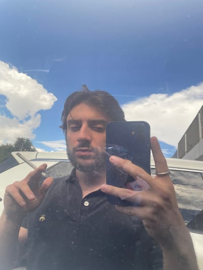 A man stands in front of a white SUV, taking a selfie with his phone. He is wearing a dark blue polo shirt with a golden logo on the left side of his chest. He is holding a lit cigarette in his left hand and has his right hand holding his phone, with a ring on his right index finger. The background shows a bright blue sky with fluffy white clouds, trees, and a building. The scene appears to be outdoors, possibly in the city of Cuautitlán Izcalli, Mexico, given the presence of an SUV and the general appearance of the surroundings.