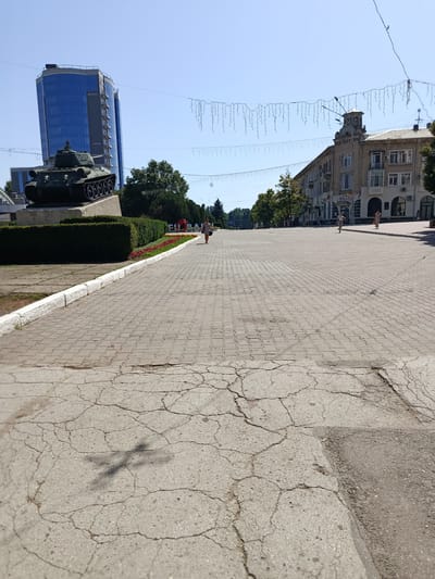The image depicts a wide, paved pedestrian area in the city of Bălți, Moldova, under a clear blue sky. A prominent feature on the left is a military tank monument situated on a raised platform, with a tall, modern glass-fronted building in the background. To the right, a row of older, multi-story buildings line the street, some with shops at ground level. Several people are visible walking on the pedestrian area, appearing to be engaged in casual movement. Strings of decorative lights are suspended across the street, suggesting a festive or public event. The foreground shows a cracked concrete surface, while the pedestrian area itself is paved with interlocking bricks. The overall environment suggests a public square or a main thoroughfare in a city setting.