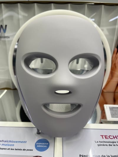 A grey LED face mask with circular eye and mouth holes dominates the image. It's surrounded by a white ring and sits atop what appears to be its packaging. Text to the right highlights the technology used, emphasizing light generation and penetration. Additional text describes it as a device for "refreshing at home," targeting skin types and tones, and is categorized as a "medical device."