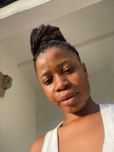 A close-up, tilted view of a Black woman with locs styled in a bun. She has a nose ring and is wearing a white top. The sunlight highlights her features, and her gaze meets the camera. A vintage telephone sits on a shelf in the background against white walls.