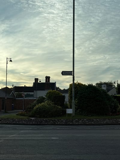 The image shows a street scene in South Staffordshire, United Kingdom, on an overcast day. In the foreground, there is a road with white lane markings. Behind the road, a low stone wall borders a grassy area with some bushes and flowers. A tall lamp post stands on the left side of the image, and another taller lamp post with a directional sign pointing to "Market" is situated more centrally. In the background, there are buildings with red brick chimneys, trees, and more street lighting. The sky is filled with textured clouds. A street sign with a no-entry symbol and another sign with "Community Cafe" and further details are visible.