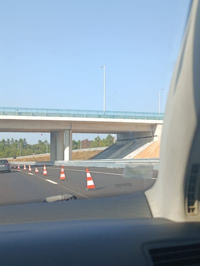 The image captures a view from inside a vehicle traveling on a highway in Santiago do Cacém, Portugal. The highway features a clear blue sky above and is lined with numerous orange and white traffic cones, indicating construction or road work. An overpass is prominently visible in the background, supported by large concrete pillars. The vehicle's dashboard and part of the interior are in the foreground, suggesting a first-person perspective of the journey. Trees and vegetation are visible beyond the highway, adding to the natural environment of the area. The overall scene depicts a segment of the highway infrastructure, likely undergoing maintenance or development, set against a sunny, outdoor environment.