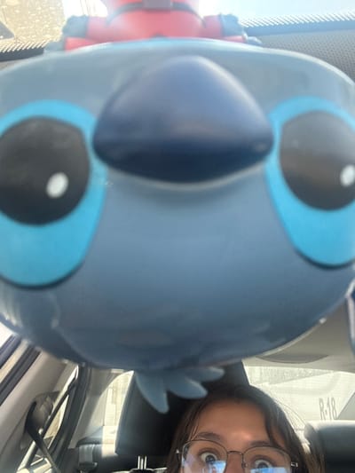 The image depicts the interior of a vehicle in Casa Blanca, Mexico, presented from a low-angle, close-up perspective. A large, light blue figurine of the character Stitch occupies the majority of the foreground, with its black eyes, white pupils, and dark blue nose clearly visible. Situated behind and above Stitch is a smaller, red and black figurine of Deadpool, visible from the chest up. At the bottom of the frame, the eyes and forehead of an individual wearing clear-rimmed glasses are visible, partially obscured by the Stitch figurine. The glasses reflect elements of the car's interior. The background beyond the figurines, viewed through the vehicle's windshield, shows bright, ambient daylight. Interior components of the car, such as a textured dashboard and a portion of a headrest, are also present.