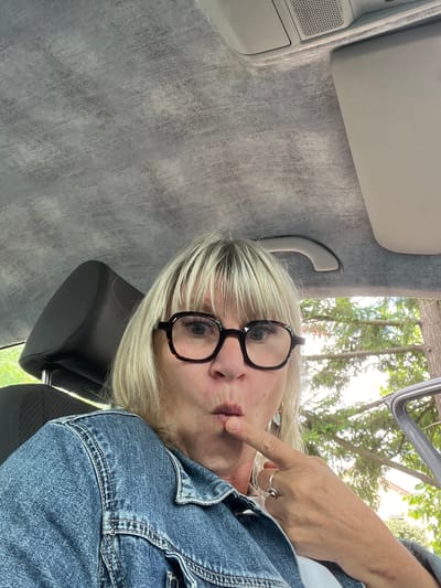 A woman is sitting in a car, looking at the camera. She is wearing glasses and a denim jacket. Her finger is on her lips, as if she is about to speak or is making a "shushing" gesture. The car's interior, including the headrest and ceiling, is visible. Outside the car, trees and a building can be seen, suggesting an outdoor setting, possibly a residential area. The lighting suggests it is daytime. This image was taken in Montrond-les-Bains, France.