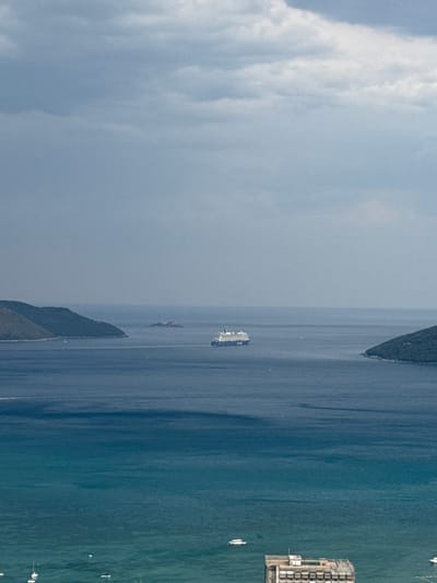 This is an aerial photograph taken from a high vantage point, likely a drone or aircraft. The image depicts a coastal scene with a body of water, possibly a bay or sea, meeting a landmass covered in lush green vegetation. Several boats are visible scattered across the water, suggesting a leisurely or commercial maritime activity. In the distance, a large white vessel, possibly a ferry or cruise ship, is navigating the water. On the right side of the frame, a complex of buildings, appearing to be industrial or resort structures, stands on the coastline, with more boats docked nearby. The water transitions in color from a deep blue to a lighter, more greenish hue closer to the shore, indicating depth variations or changes in water composition. The sky is partly cloudy, with soft, diffused light suggesting it is daytime, possibly late morning or afternoon. The scene conveys a sense of calm and expanse, with the natural beauty of the coastline juxtaposed with man-made structures and vessels. The overall impression is of a serene, active coastal environment.