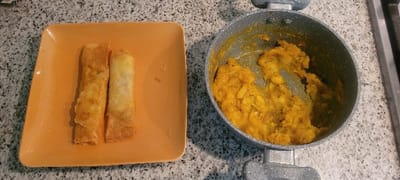 An orange square plate holds two spring rolls, one golden brown and slightly crumpled, the other lighter in color. Adjacent to the plate, a pot on a stove top contains a yellow, mashed mixture, possibly a filling for the spring rolls. The countertop is a speckled gray and white. A section of a black stove element is visible to the right of the pot.