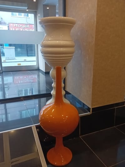 The image depicts an indoor scene with two decorative vases. The larger vase, predominantly white, has a ribbed texture and a wide base, tapering upwards to a rounded opening. To its right, a second vase, bright orange, stands on a pedestal. This orange vase features a bulbous lower section connected to a slender neck and a flared opening. Both vases are positioned on a dark, reflective floor. In the background, through a glass storefront, glimpses of an urban environment are visible, including buildings and signage. One sign, reflected in the floor, appears to read "Royal İletişim." The overall impression is of an interior space likely a shop or establishment in Kırıkkale, Turkey, showcasing decorative items.
