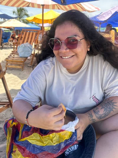 A female subject is depicted from the waist up, seated on a sandy beach in General Villamil Playas, Ecuador. She has dark, long, wavy hair and is wearing hexagonal, pink-tinted sunglasses and a light-colored, short-sleeved t-shirt with a small pink centipede graphic on the left chest. Her left forearm displays a prominent tattoo featuring a sun and moon motif with additional script. A black beaded bracelet and a red string bracelet are visible on her left wrist. She smiles directly at the camera. Her right hand holds a piece of light-colored food, possibly bread, over an open container wrapped in a white plastic bag, which is held in her left hand. A multicolored plastic bag (red, yellow, blue stripes) rests beside her left side, partially obscuring the container, and a dark bucket hat is visible near her left leg. The background features a vibrant beach scene with multiple colorful umbrellas (striped orange/white, yellow, blue, purple) providing shade, along with several wooden beach loungers. Other indistinct figures are present in the distance. The setting suggests clear, bright daylight.