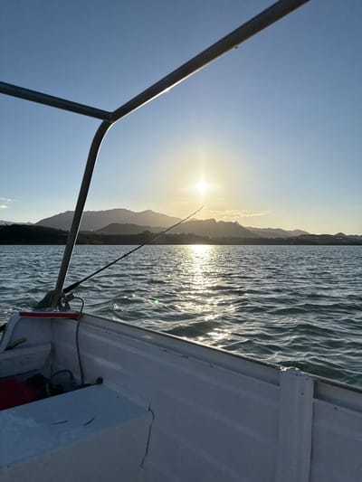 The scene depicts a small, white, open-top boat positioned on a body of water in Dumbéa, New Caledonia, France, during either sunrise or sunset. The perspective is from within the boat, looking forward towards the horizon. The sun, appearing as a bright, star-like point of light, is situated low in the sky, slightly to the right of the image center, casting a prominent, shimmering reflection path across the rippled water. A dark-colored fishing rod extends from the port (left) side of the boat, angled upwards and towards the sun. In the distant background, a range of dark, silhouetted mountains forms a landmass, with faint indications of structures at their base on the right side. The sky above is clear and light blue. Within the boat, white storage compartments or benches are visible, along with a partially obscured red object and a coiled rope in the bow area. A metallic arch frame, part of the boat's structure, extends from the left side into the upper left of the frame. No individuals are present in the image.