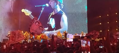 This image depicts a vibrant live music concert at night in Tolosa, Argentina. The central focus is a band performing on a brightly lit stage, backed by a massive LED screen that displays a close-up of the lead singer. The singer, a man wearing a baseball cap and a dark sleeveless shirt, is passionately singing into a microphone while playing an electric guitar, whose Fender-style headstock is clearly visible.
Other band members are visible on stage, including a drummer behind a drum kit and additional musicians with guitars. A distinctive stage prop on the left features a large, sculptural element resembling a monster or skeleton's head, from which a giant electric guitar headstock emerges. In the foreground, a large crowd of concert-goers is actively engaged, with many hands raised in excitement and numerous smartphones held aloft to record the event. Several attendees are waving red banners or flags; one on the left prominently displays the text "LA GENTE QUE SIEMPRE" (THE PEOPLE WHO ALWAYS). A black circular object on stage, possibly a drumhead or speaker, shows an infinity symbol (∞). The overall atmosphere is energetic and enthusiastic, illuminated by stage lighting against the dark backdrop.
