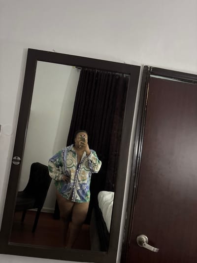 A mirror reflects a woman wearing a patterned shirt, appearing to take a selfie. Behind her, a dark curtain is visible. The setting includes a wooden door above the mirror and a chair within the room's reflection. It is not possible to determine if the location is in Port Harcourt, Nigeria from the image.