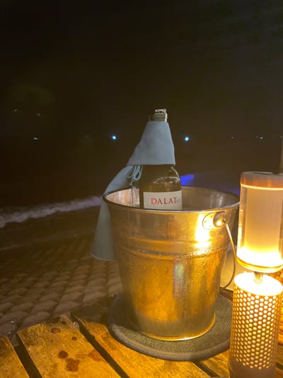 The image depicts a nighttime scene in Thành phố Phan Thiết, Vietnam, featuring a beverage setup on a wooden table at the edge of a body of water.
In the foreground, a dark-stained, slatted wooden table surface is illuminated by a warm, golden light source originating from the right. On this table, slightly left of center, rests a metallic, brass-colored ice bucket, visibly covered in condensation. The bucket is placed on a dark, circular woven coaster. Inside the bucket, a dark-colored wine bottle stands upright, its label, partially visible, reads "DALAT". A light blue fabric cloth is draped over the neck and shoulder of the bottle, with the bottle's corked top, encased in foil, extending above the fabric.
To the right of the ice bucket, a two-tiered table lamp is present. The lower section of the lamp is a cylindrical, perforated base emitting warm light, while the upper section is a translucent cylinder, also emitting warm light.
The background reveals a dark night sky. To the left, the dark surface of an ocean or large body of water is visible, with faint white lines indicating small waves breaking near the shore, illuminated by ambient light. Several small, distant blue lights are discernible across the dark expanse of the water in the mid-background. No individuals are present in the scene.