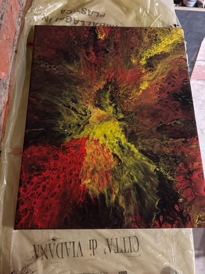 The image shows a close-up, overhead view of an abstract acrylic pour painting with vibrant colors. The painting is primarily composed of swirling patterns of red, yellow, and black. The colors are blended in a way that creates a dynamic, explosive effect, with a bright yellow center radiating outwards into fiery reds and dark blacks. The painting appears to be on a canvas and is resting on a light-colored plastic sheet that has text printed on it. The text is in Italian and appears to read "CITTA' di VIADANA" and other fragmented words. The background is out of focus, but glimpses of a brick wall on the left and a dark surface on the right suggest an indoor or covered outdoor setting. There are no people or animals visible in the image. The overall impression is one of intense energy and movement captured in paint. There is no indication of Villa Pasquali, Italy in the image.