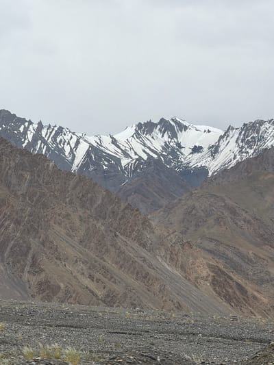 A wide shot captures a rugged, mountainous landscape with a rocky, desolate foreground. In the distance, towering peaks are capped with snow, their jagged ridges contrasting with the muted, cloudy sky. The middle ground features steep, rocky slopes exhibiting a layered geological structure, with shades of brown and grey dominating the terrain. Patches of sparse, dry vegetation are scattered across the rocky foreground, adding a touch of muted color to the otherwise barren scene. The overall impression is one of an isolated and harsh natural environment.