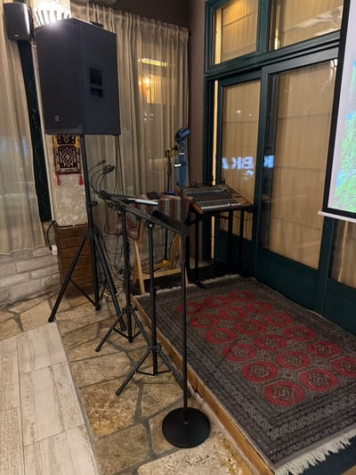 An indoor setup for a performance is depicted. A large black speaker is mounted on a stand on the left. In the center, a mixing board sits on a stand, with multiple microphones positioned around it. A projection screen is visible on the far right, partially obscured by a glass-paneled door. The floor is a mix of stone and tiled surfaces, with a patterned rug placed in front of the mixing board. The environment appears to be a dimly lit interior space, possibly a cafe or a small venue, with curtains and lighting visible in the background. There are no people present in the image. The location is identified as Kumbor, Montenegro.