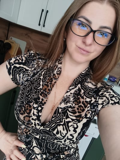 A woman with long brown hair, wearing black-framed glasses, a patterned dress with a V-neck, and a gold necklace with a cross pendant, is posing for a selfie indoors. She has her left hand on her waist. The background features kitchen cabinets in white and green, along with kitchen utensils on the counter. The specific location within the city of Chernogolovka, Russia is not discernible from the image.