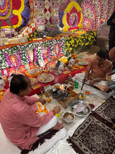 An indoor scene in Bidhannagar, India, depicts a Hindu ritual (puja/havan) in progress. Three adult males are seated on white floor coverings over patterned brown rugs.

The background features an elaborately decorated wall. This backdrop includes large floral arrangements in yellow, red, and purple hues, intertwined with patterned fabric panels (red, gold, purple). A shelf-like structure, also adorned with green foliage and flowers, displays numerous small framed images, likely of deities, and various packaged items. White spotlights illuminate this area.

In the midground, a red cloth covers a table holding an extensive array of offerings. These include fresh fruits (oranges, pomegranates), numerous packaged sweets in boxes and trays, and small metal bowls containing various powders or liquids.

In the foreground, the ritual is centered around a square metal brazier containing burning embers, from which smoke rises. Several small metal bowls containing colored powders (e.g., red, orange, yellow) and liquids are arranged around the brazier. A larger metal bowl holds an assortment of Indian currency notes. Another small metal bowl contains unidentifiable ritual items.

A male individual on the left, with a receding hairline and wearing a light pink kurta and white trousers, is seated cross-legged, facing right. A yellow and white striped cloth rests on his lap. He holds a small orange object in his right hand.

A second male individual, center-right, wearing a patterned orange and white short-sleeved kurta and white trousers, is seated cross-legged. He holds a long, dark instrument with a spoon-like end, seemingly manipulating items within a small bowl near the brazier.

A third male individual is partially visible on the far right, with a bald head and wearing a light-colored patterned shirt. He is holding a small, rectangular, packaged item in both hands.