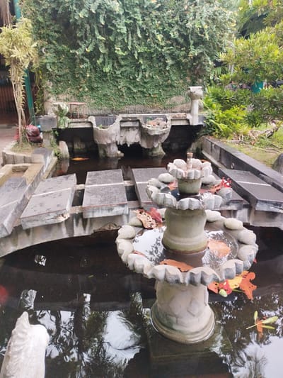 In Payameuneng, Indonesia, a stone fountain stands in the center of a dark water pond, surrounded by concrete platforms and lush greenery. The fountain has two tiers, with the upper tier overflowing onto the lower one. Fallen leaves, some bright orange and yellow, float on the water. The pond is bordered by grey concrete structures, some with a textured surface, and what appear to be submerged stepping stones. Behind the pond, a wall is heavily covered in green vines, creating a natural backdrop. To the left of the pond, a tall, thin tree with light-colored leaves grows near a pile of red bags. To the right, more dense green foliage and trees are visible. The water reflects the surrounding environment, creating a somewhat distorted image of the greenery and sky.