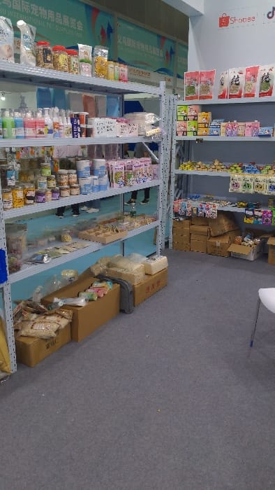 The image shows a section of a large retail space, likely a trade show or convention hall, located at 30.319 degrees latitude and 120.357 degrees longitude in Hangzhou Shi, China. The scene contains four rows of shelves stacked with various pet products, including packaged food, grooming supplies, and toys. In the foreground, there are boxes of pet supplies and a white chair. In the background, there is a banner with Chinese writing. The shelves are made of metal and have a light blue finish. There are no people visible in the image. The floor is a light gray carpet. The lighting in the space is bright and artificial. The air is clean and well-lit. The overall tone is one of sterile commercialism.