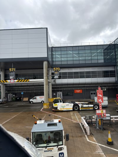 This image presents a forensic view of an airport apron and terminal building, specifically depicting ground operations at London Gatwick Airport, located near Crawley, United Kingdom. The perspective is from an elevated position, likely an aircraft window or an upper level of the terminal, looking down onto the tarmac and adjacent structures.
The main terminal building is constructed with modern grey and white paneling and extensive dark-framed glass windows. Prominently displayed are two gate numbers in yellow: "571" to the left and "570" to the right. Below gate "571," a yellow and black striped height restriction bar is visible, marked "4.2 m 13'9"". Further below, a blue arrow sign points left towards a service area.
On the wet concrete apron, several ground support vehicles and equipment are present. A white and yellow aircraft tow tractor, bearing "PHOENIX E" and "ASTRE" branding with a green leaf logo, and a visible partial number "937," is parked centrally. To its right, a red and white warning sign indicates "Emergency Stop Buttons," "Fuel Hydrant," and "Aircraft," with an arrow pointing downwards. Adjacent to this, another red and white sign displays symbols for aircraft and segregation, with text including "SEG 571," "SEG 570," and "SEG 575." A stack of yellow traffic cones rests near a white railing and utility boxes.
Further left, a white van marked "easyJet Engineering" is parked near green waste bins and a wooden pallet. In the immediate foreground, the top portion of another ground support vehicle, labeled "TREPEL" and "RUSHLIFT" with the number "609832," is partially visible. The entire scene is under an overcast sky, characterized by uniform grey clouds, and the ground appears damp from recent precipitation. No individuals are clearly visible.