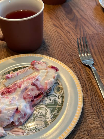 The image shows a slice of dessert on a decorative plate, a fork, and a mug of dark liquid, likely tea, on a wooden table. The dessert appears to be a layered confection with white cream and red fruit filling. The plate has a pale green rim with a yellow and white floral pattern. The mug is a dark reddish-brown color and is partially visible in the upper left corner. The fork is made of metal and is positioned to the right of the plate. The wooden table has a visible grain pattern. There are no people or actions depicted. The image is a still life of food and drink on a table, with no discernible location in Shepelyovo, Russia.
